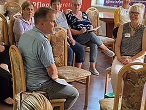 Großer Stuhlkreis in einem Raum mit Kachelofen. In der Mitte ein kleiner Stuhlkreis 5 Stühle. Ca. 30 Personen im Raum. Viele ältere Frauen.