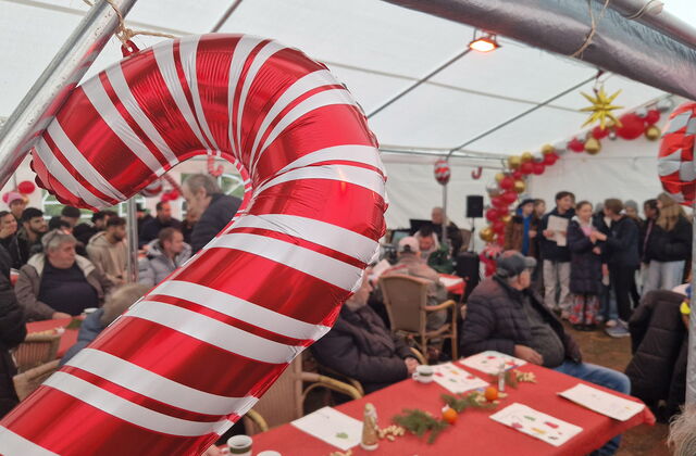 Im inneren des großen Festzeltes. Tische Stühle, viele Menschen. Der Chor steht vorn und singt. Im Vordergrund des Bildes ist ein Luftbalon in Form ein rot-weiß gestreiften Zuckerstange.
