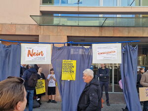 Draußen auf dem Gehweg vor einem großen Gebäude. Eine blauer Vorhang, daran sind Plakate geheftet: Gelbes Plakat: Demo gegen Kürzungen.