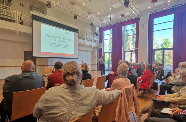 Großer Saal. Viele Stuhlreihen wo Menschen sitzen, Leinwand forne, ein Redner mit Mikrofon steht vorn.