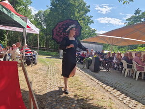 Eine Frau präsentiert Kleidung auf einer Modenschau. Frau trägt ein schwarzes knielanges Kleid und einen dunklen Sonnenschirm als Accessoires