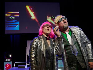 Theaterstück. Zwei Persoonen auf der Bühne. Einne Frau mit pinker Perücke und einer schwarz-roten Brille sowie Fliege in schwarz gekleidet. Der Mann blond mit grün-scharzer Brille, grünem T-Shirt, weißer Krawarre hält einen kleinen Bann hoch.