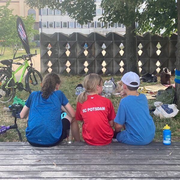 Draußen, Sonne, schattenspendender Baum. Drei Kinder sitzen auf etwas aus Holz. Unter dem Baum ist eine Decke ausgebreitet und Rucksäcke und Taschen liegen rum. Die Kinder tragen T-Shirt. Mädchen in der Mitte ein rotes T-Shirt vom SC Potsdam. Von hinten,