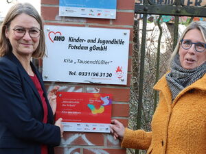 2 Frauen stehen vor einem Schild der Kita Tausendfüßler. Sie zeigen auf eine Roto Plakette darunter "Auszeichnung Kita des Jahres".