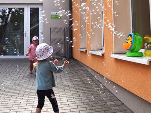 Eine Seifennblasenmaschine steht auf einem Fensterbrett und macht viele Seifenblasen. Zwei kleine Kinder erfreuen sich daran und versuchen die Seifenblasen zu fangen.