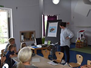 Ein Raum. An einer Tafel ein Bild, daneben steht ein Frau die erklärt. An einem Tisch sitzen zwei Kinder Junge und Mädchen und Frauen.