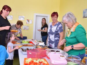 Ein gelber Raum. Darin ist das Büfett: Gurkenscheiben, Tomaten, Kuchen, Muffins, Trinkpäckchen. Zwei ältere Frauen verteilen das Gewünschte an Kinder und Erwachsene.