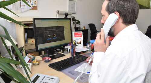 Ein Mann in weißem Hemd sitzt in einem NBüro am Schreibtisch, vor ihhm der Monitor, Tastatur unnd ein Notizblock, wo er etwas notiert, er hat den Telefonhörer am Ohr.