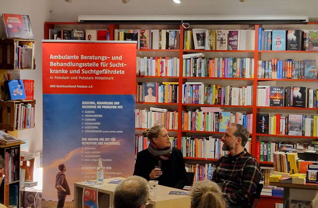 Eine Frau und ein Mann sitzen an einem Tisch und sehen sich an. Neben dem Tisch ein Roll-Up der AWO Suchtberatung.       Hinter ihnen große Bücherregale. Publikum sitzt im Raum.