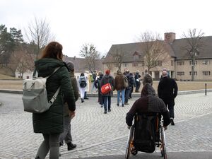 Mehrere Menschen laufen auf ein Gebäude zu, ein Rollstuhlfahrer ist auch darunter. Alle von hinten. Der Himmel ist grau und die bäume sind kahl.