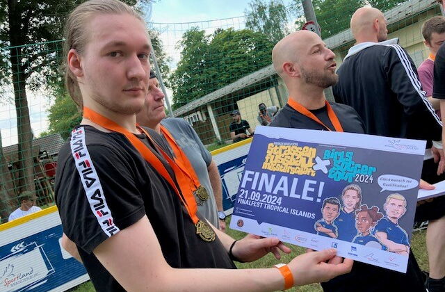 Junger Mann hält den ersten Preis in den Händen, er ist blond und hat eine Medaille an einem orangen Band um den Hals hängen. Preis: große Karte Text: "Straßenfußball. Finale 21.09.2024 Finalfest Tropical Island". Zwei weitere Spieler mit Medaille im Bild