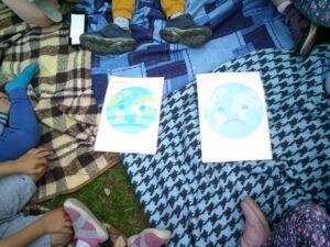 Zwei Bilder der Erde. Eine lachende Erde/Welt und eine traurige Erde/Welt auf Decken. Kinder sitzen drumherum.