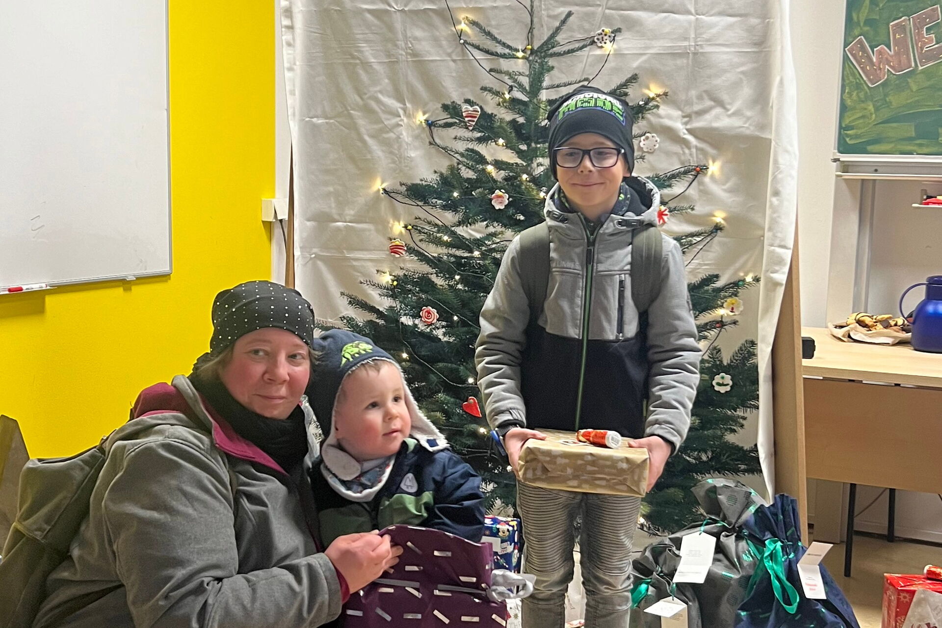 Geschenkeübergabe. Klassenraum. Frau hockt bei einem kleinen Kind, das Kind hat ein großes Geschenk. Vor einem kleinen Weihnachtsbaum, steht ein Junge auch mit Geschenk in den Händen.