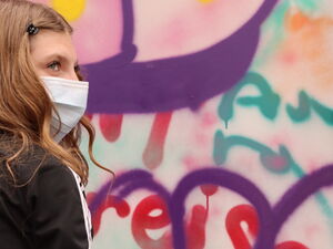 Frau mit weißen Handschuhen und OP-Maske, Sie greift nach eine Spraydose. Vor ihr eine besprühte Graffiti Wand.