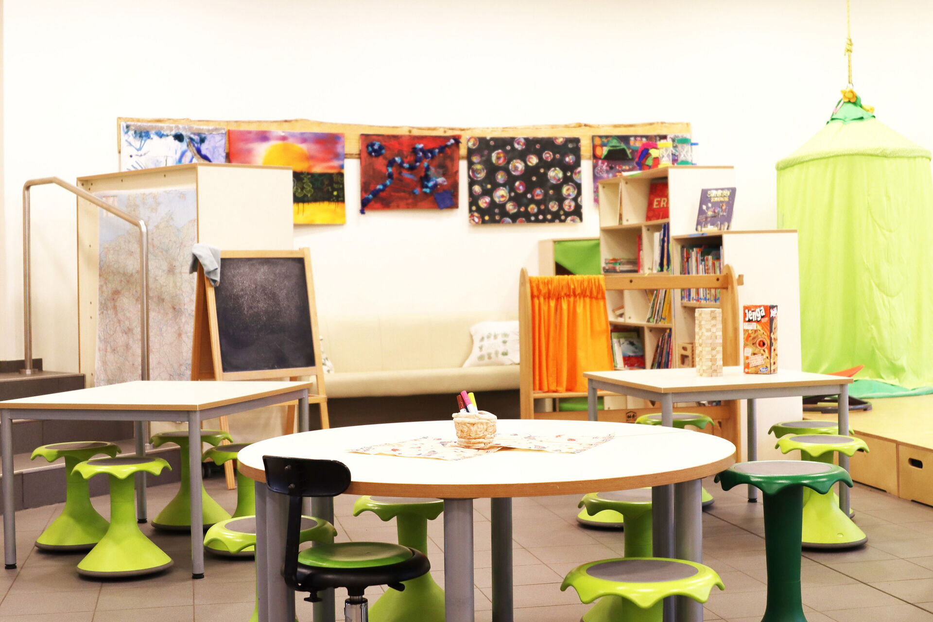 Ein Raum mit drei kleinen Tischen mit je 4 Hockern. Bilder hängen an der Wand. Kindertafel, grüne Hängehöhle und ein kleines Regal mit Büchern.