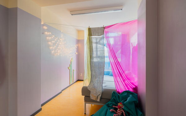 Kleiner Raum, mit leuchtender Lichterkette an der Wand. Farbige Tücher hängen vor einer Liegefläche. Durch ein Fenster fällt Licht in den Raum.