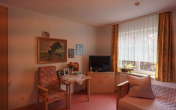 Zimmer. Großes Fenster rechts, Fernseher auf Kommode in der Ecke, kleiner Tisch und Stuhl daneben. Über dem Tisch ein Landschaftsbild.