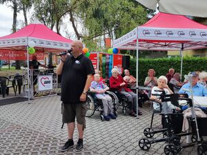 Draußen. Senioren sitzen in Rollstühlen und auf Stühlen an Tischen. Zwei AWO Pavillons.Ein kräftiger Mann mit Glatze spricht in ein Mikrofon. Mann trägt schwarzes T-Shirt und Kaki kurze Hose.