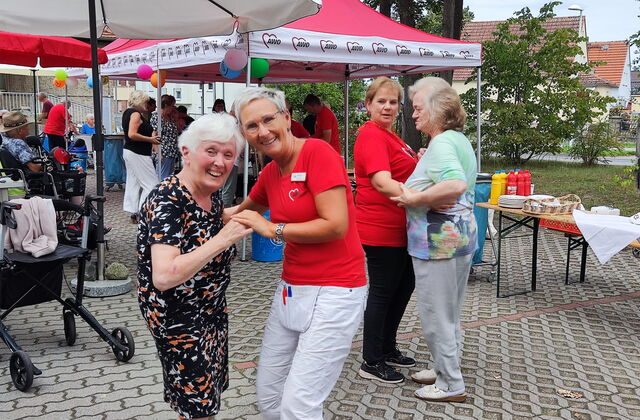 Zwei Helferinnen in roten AWO T-Shirts tanzen fröhlich mit zwei alten Damen. Die Tanzenden im Vordergrund lächeln in die Kamera.