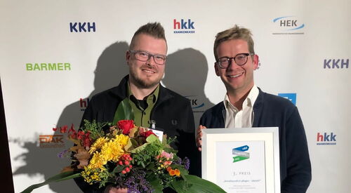 Zwei Männer vor einer Werbewand. Der jüngere Mann hat einem großen Blumenstrauß in der rechten Hand. Der Mann daneben hält eine gerahmte Urkunde zum 3. Platz vor sich.