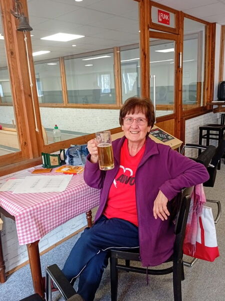 Frau mit rotem AWO T-Shirt und lila Strickjacke sitzt im Aufenthaltsraum mit Bierglas in der Hand.