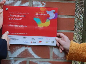2 Frauen stehen vor einem Schild der Kita Tausendfüßler. Sie zeigen auf eine Roto Plakette darunter "Auszeichnung Kita des Jahres".