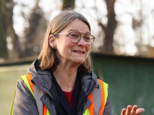 Frau mit grauer Jacke, Neon-farbiger Weste und Brille streckt eine Hand nach vorn.