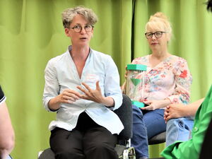 Zwei Frauen sitzen auf Stühlen, vor einem grünen Vorhang. Die eine Frau sitzt im inneren Kreis und spricht. Sie hält eine Hand vor sich mit der andere zählt sie etwas an den Fingern ab. Die Frau die hinter ihr sitzt hat eine grüne Sanduhr auf dem Schoß.