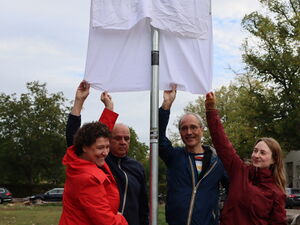 Ein Straßenschild mit einem weißen Tusch darüber. Von unten greifen 2 Frauen und 2 Männer nach dem Tusch. Die Enthüllung steht kurz bevor.