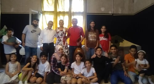 Gruppenfoto im dunklen Raum: Erwachsene und Kinder unterschiedlicher Nationen haben sich versammelt. Die Kinder sitzen und knien vorne, Erwachsene und Jugendliche stehen dahinter.