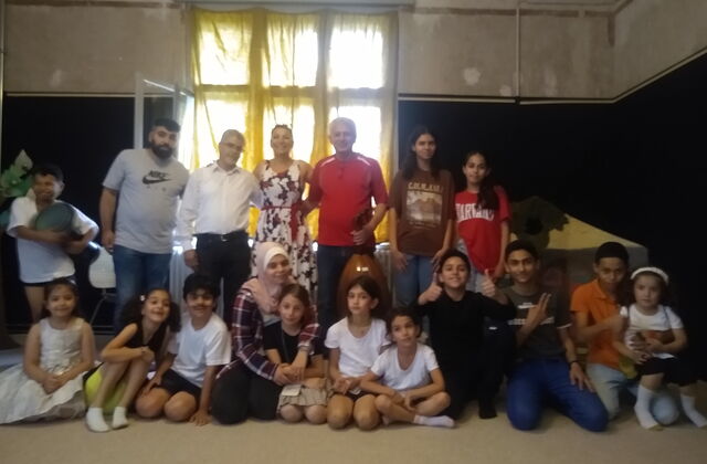 Gruppenfoto im dunklen Raum: Erwachsene und Kinder unterschiedlicher Nationen haben sich versammelt. Die Kinder sitzen und knien vorne, Erwachsene und Jugendliche stehen dahinter.