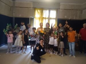 Gruppenbild. Kinder und Jugendlich zeigen den Damen hoch. In der Mitte steht Frau mit weiß-schwarzem Kleid und Blumenstrauß in der Hand. Menschen aus verschiedenen Nationen.