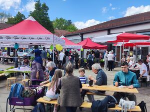 Draußen, blauer Himmel. Drei AWO-Pavillons und zwei Sonnenschirme, viele Menschen die an Biertischgarnituren sitzen, etwas essen oder sich auf dem Gelände umschauen.