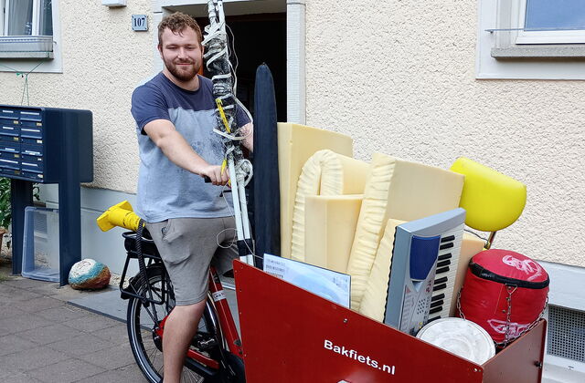 Mann mit T-Shirt und kurzer Hause sitzt auf Lastenfahrrad, der Laderaum ist voll beladen.