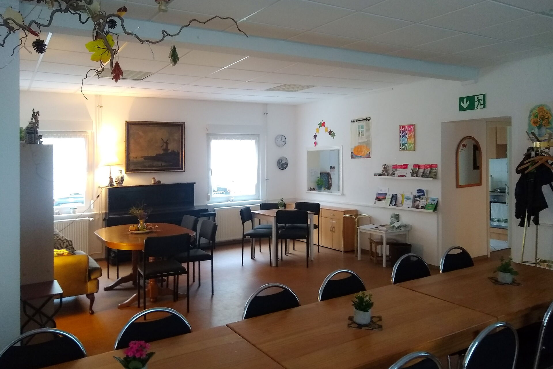 Großer Raum mit Tischen und Stühlen. Eine lange Tafel im Vordergrund und kleine Sitzgruppen sowie ein Klavier im hinteren Bereich.