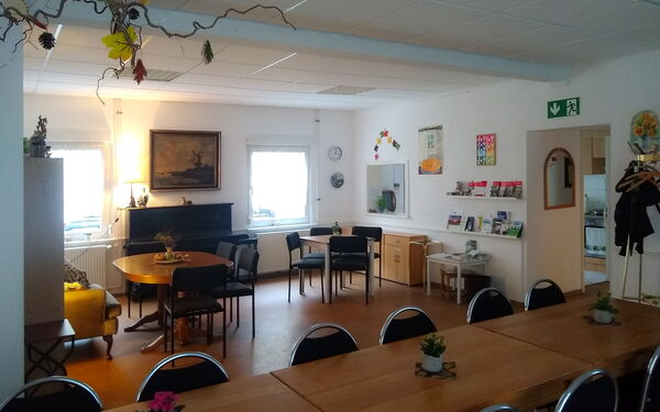 Großer Raum mit Tischen und Stühlen. Eine lange Tafel im Vordergrund und kleine Sitzgruppen sowie ein Klavier im hinteren Bereich.