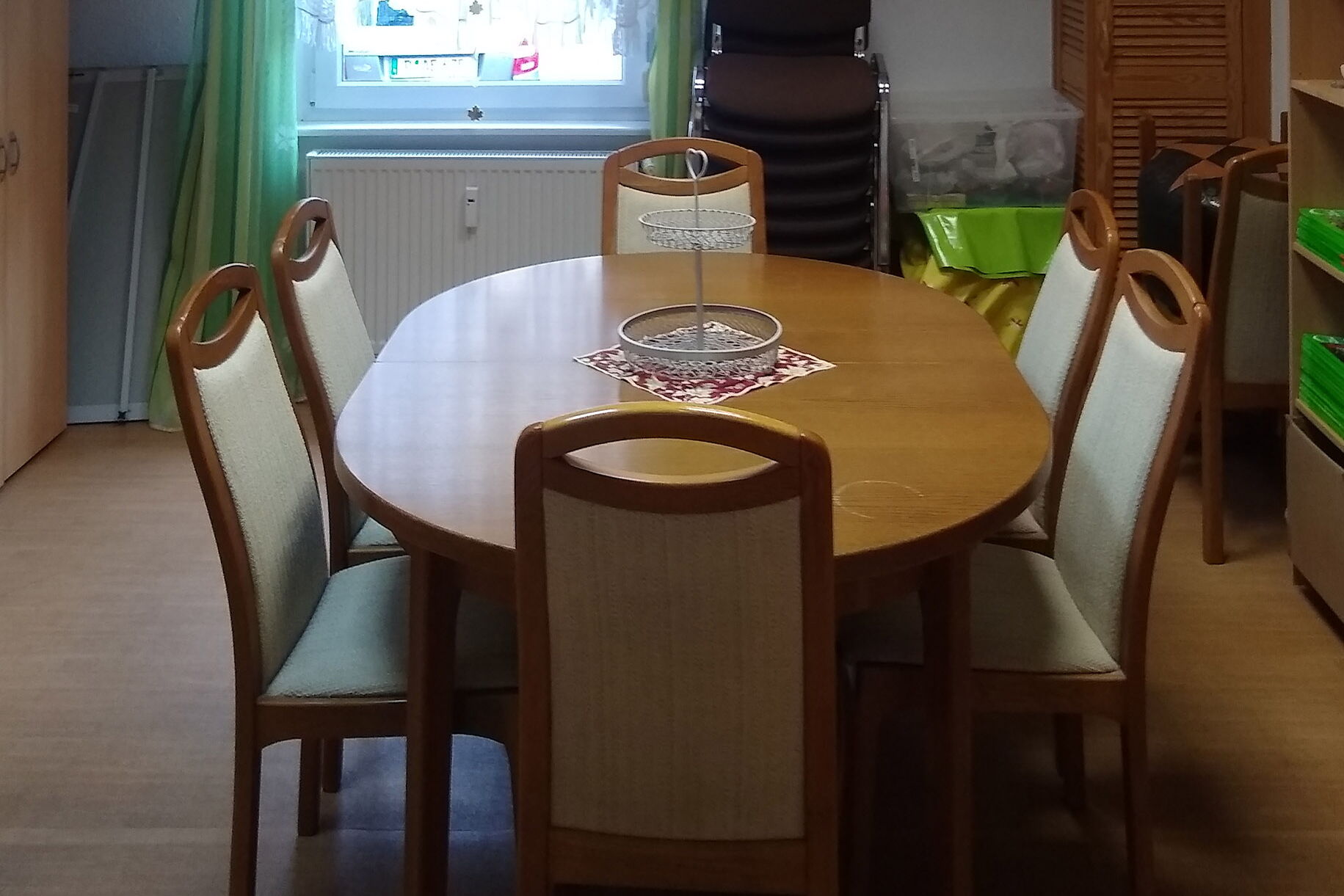Ein ovaler Holztisch und sechs gepolsterten Holzstühlen. Im Hintergrund ein Fenster mit grün-weißen Vorhängen.