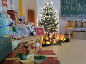 Viele Geschenke auf Tischen, ein Weihnachtsbaum, mit Lichterkette die leuchtet. Unter den Tannenbaum Geschenktüten. An einer Schultafel steht frohe Weihnachten.