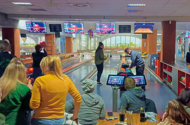 Eine Bowlinghalle. Es wird gebowlt, einige sitzen am Tisch mit Getränken und schauen zu.