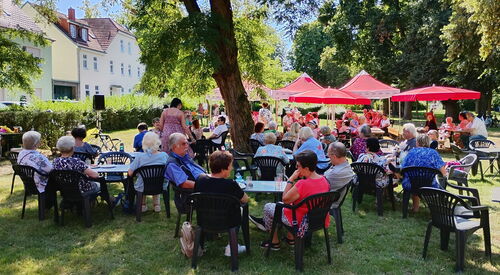 Große Wiese mit Schattenplätzen unter Bäumen und roten Sonnenschirmen. Viele Menschen sitzen draußen.