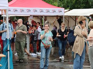 Viele Menschen bei einem fest, es hat geregnet, es sind Pfützen auf den Wegen. Menschen mit Essen und Getränken, es ist ein fröhliches Beisammensein.