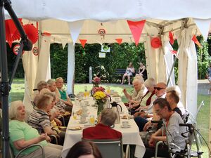 Draußen, Festzelt, mit rot-weißen Wimpel, roten und weißen Luftballons geschmückt. Lange Festtafel an der Senioren mit Kaffee und Kuchen sitzen, Das Wetter ist sonnig.