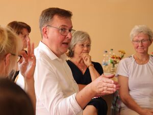 Nahaufnahme. Mann hat das Wort. Er zeigt mit der rechten Hand in die rechte Richtung. Mann trägt ein weißes Hemd mit Kragen, eine schmale Brille, die Haare sind kurz. Hinter ihm im zweiten Stuhlkreis sitzen Frauen.