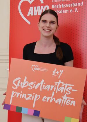Portrait: Junge Frau, mit langen braunen Haaren zum Pferdeschanz gebunden, mit schwarzem T-Shirt und heller Hose. Sie hält ein großes Schild vor sich. Eine der 1+9 Forderungen. Darauf steht: Subsidiaritätsprinzip erhalten.