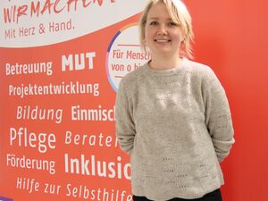 Portrait: Junge blonde Frau lächelt in die Kamera. Sie trägt einen beigen Pullover, eine schwarze Hose und hat die Hände hinter dem Rücken.