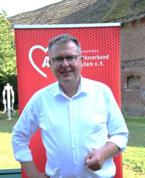 Portrait: Mann lächelt. Er hat kurze Haare, Brille und trägt ein weißes Hemd. Er steht draußen vor einem roten AWO Roll-Up-Banner