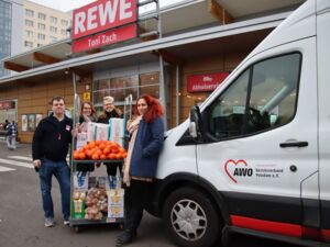 Vor einem REWE Markt. Ein weißer Transporter der AWO steht davor. Eine Frau mit langen wilden roten Haaren lehnt am Transporter. 3 weitere Personen schieben einen Rollbehälter beladen mit Lebensmitteln. Alle lächeln in die Kamera.