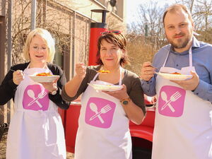 2 Frauen und ein Mann mit weißen Schürzen und Tellern in der Hand. Sie lächeln in die Kamera.