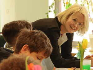 Kinder beim gemeinsamen Essen am Tisch. Am Tischende steht eine blonde Frau mit Brille die sich auf den Tisch lehnt und zur Kamera lächelt.