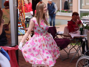Eine junge Frau in kurzem Sommerkleid mit pinken Blumen und weißen Handschuhen macht einen Knicks.